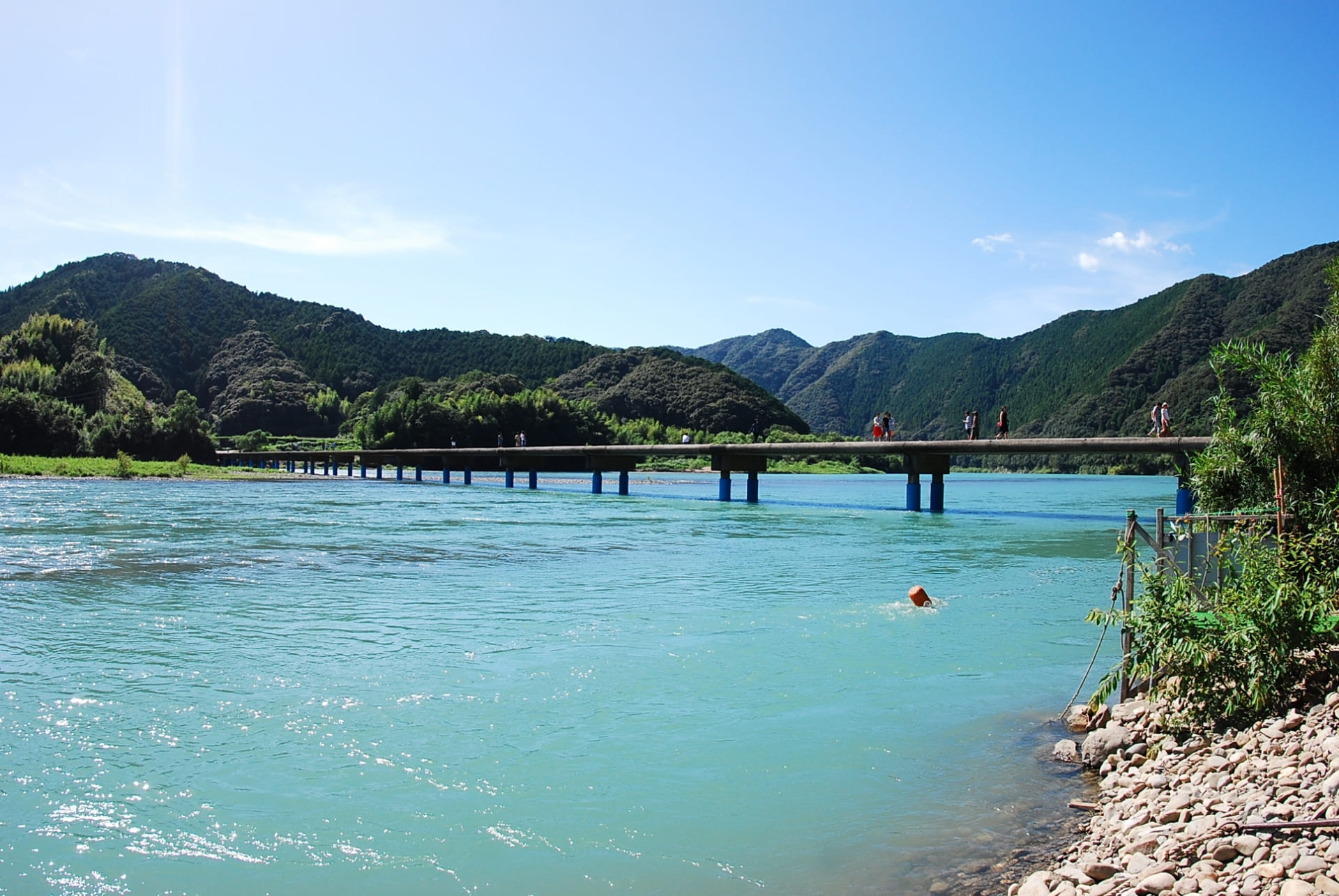 夏の四万十川に架かる沈下橋。青空と緑の山を背景に、欄干のない橋を人々が渡っている