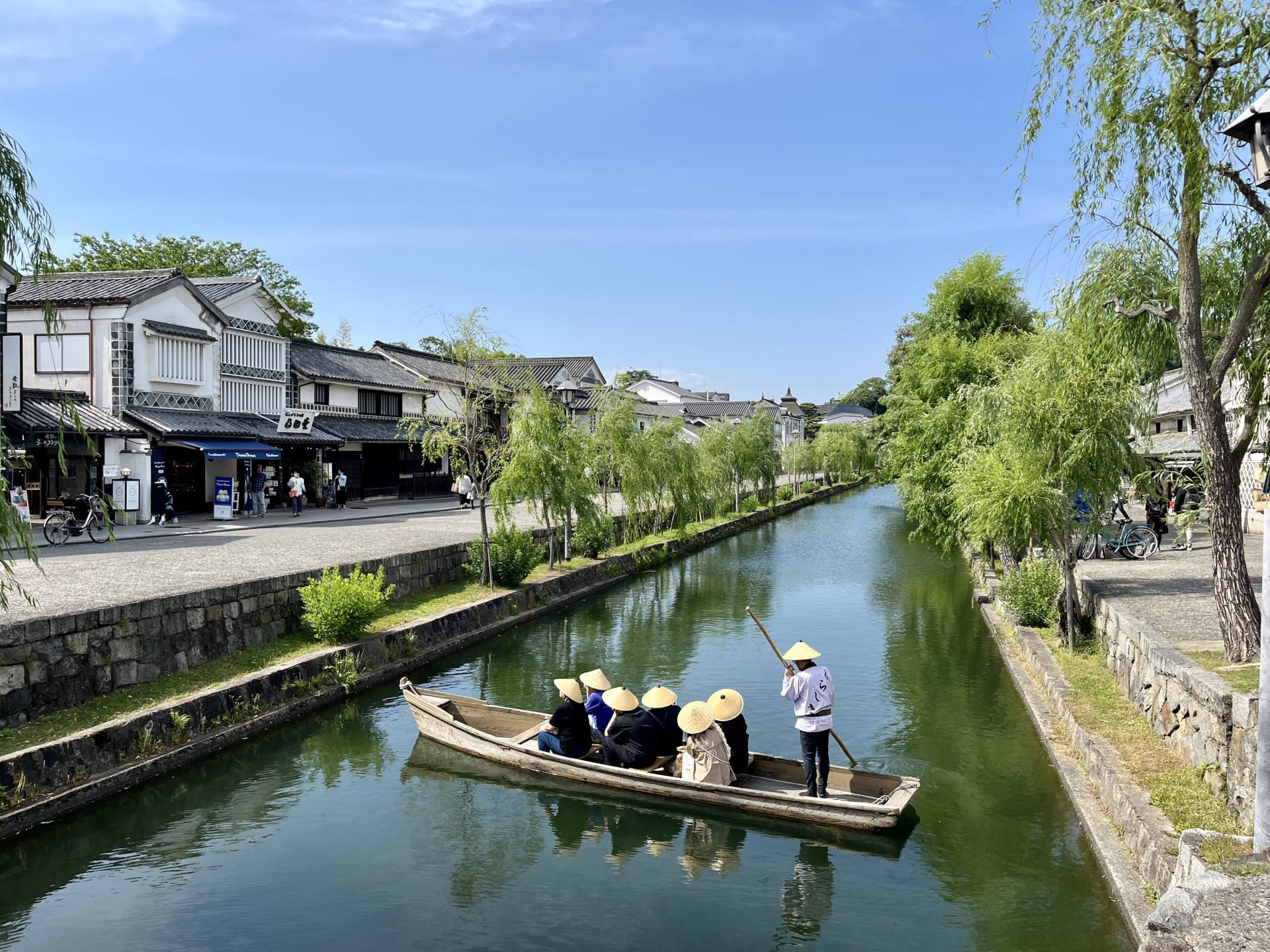 晴れた日の倉敷川を進む川舟。船頭が竿で舟を操り、柳並木と白壁の蔵屋敷が両岸に連なる